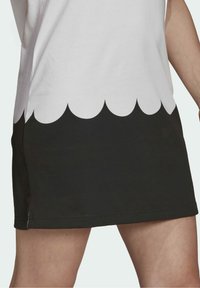 Jupe blanche et noire avec un bord festonné portée par une personne, montrant une longueur mi-cuisse et une partie des jambes et des bras nus.