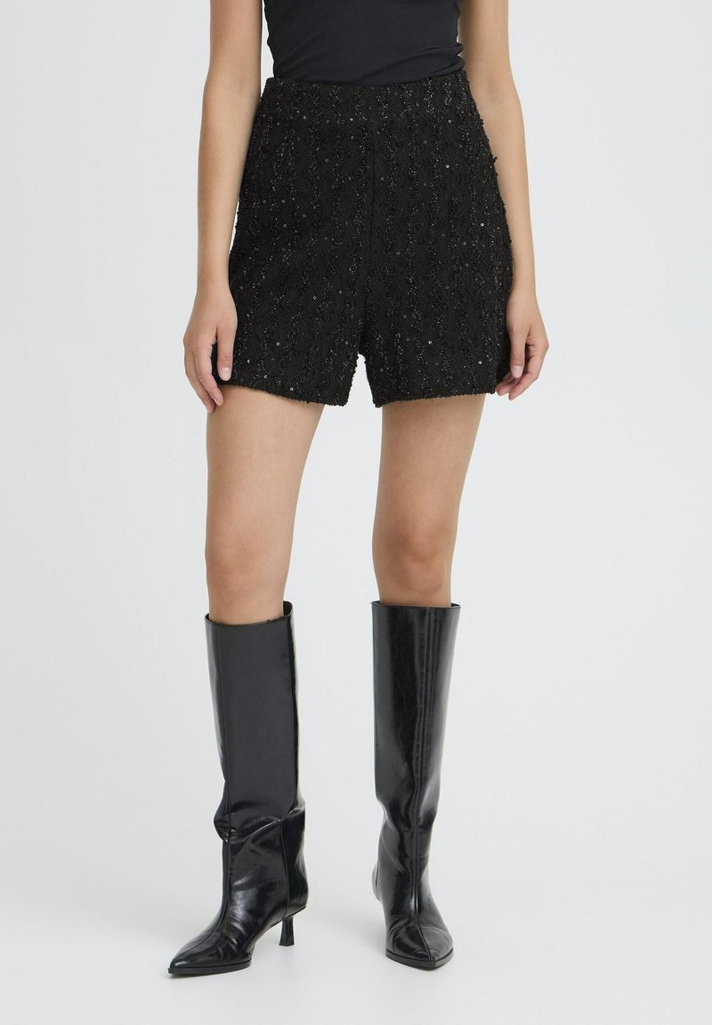 Femme portant un short noir texturé taille haute associé à des bottes en cuir noir brillantes montantes jusqu'au genou et un haut noir sur un fond uni.