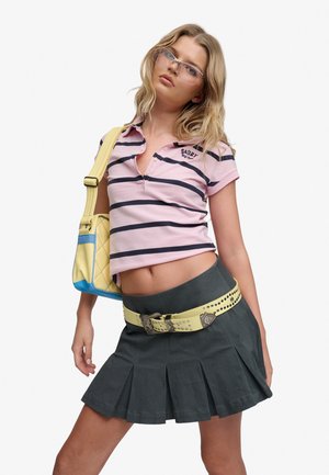 Jeune femme portant des lunettes transparentes, une chemise rose à rayures, une jupe plissée foncée avec une ceinture cloutée jaune, et portant un sac à main jaune avec des accents bleus.