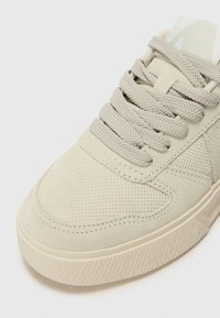 Sneaker beige con tomaia in suede testurizzata, lacci piatti e suola in gomma a motivo. Presenta un design minimalista e una palette di colori chiari.