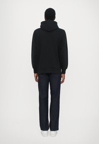 Black hooded sweatshirt with a relaxed fit, paired with dark blue jeans and white sneakers, seen from the back.