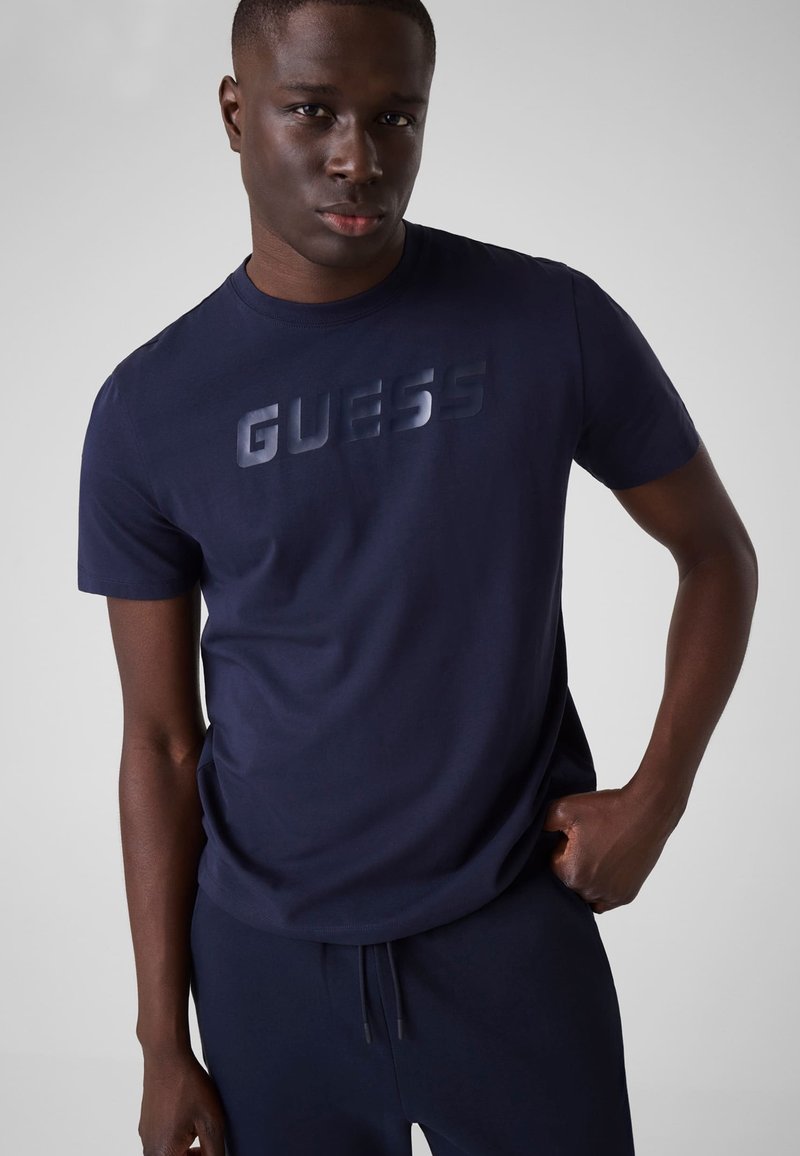 Άνδρας με μπλε σκούρο μπλουζάκι GUESS και αντίστοιχο παντελόνι στέκεται με το ένα χέρι στην τσέπη, κοιτώντας μπροστά σε απλό φόντο.