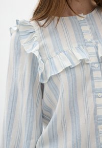 Blusa a righe blu chiaro e bianca con scollo arricciato, chiusura a bottone e tessuto testurizzato. Presenta righe verticali e morbide ondulazioni.