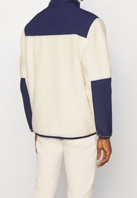 Fleecová bunda v krémové barvě s tmavě modrými panely. Má vysoký límec a kontrastní texturovanou látku. Zobrazeno zezadu, s bočními kapsami.