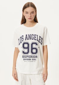 T-shirt en coton blanc avec col rond, présentant un imprimé graphique bleu marine : "LOS ANGELES 96 DIVISION SUPÉRIEURE 0301 CALIFORNIE" en lettres gras.
