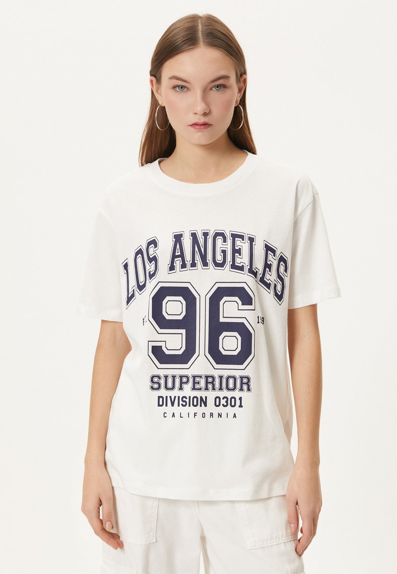 T-shirt en coton blanc avec col rond, présentant un imprimé graphique bleu marine : "LOS ANGELES 96 DIVISION SUPÉRIEURE 0301 CALIFORNIE" en lettres gras.