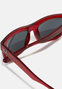 Røde plastik solbriller med mørkegrå linser, der har et skarpt, vinklet design og tykke stel. Ray-Ban-logoet er synligt på siden.