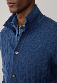 Blauwe kabelgebreide trui met hoge kraag, voorzien van een geribd ontwerp en een zwarte knoopsluiting, gedragen over een lichtblauw overhemd met kraag.