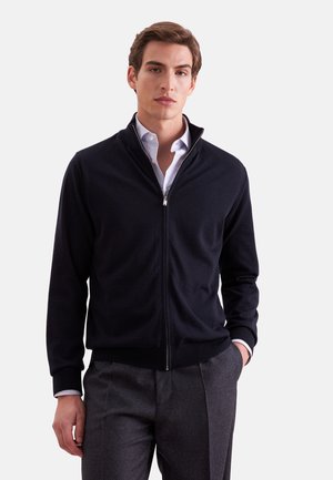 Junger Mann trägt einen schwarzen Zip-up-Pullover über einem weißen Hemd und grauen Hosen, steht mit einer Hand in der Tasche vor einem einfarbigen Hintergrund.