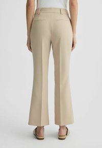 Pantaloni beige a gamba larga con una texture morbida, dettagli sulla tasca posteriore e fascia in vita sagomata. Abbinati a scarpe beige senza lacci.