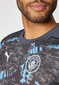 Black graphic t-shirt with blue accents, featuring a Puma logo and Manchester City emblem on the chest. Made of soft fabric with textured design.