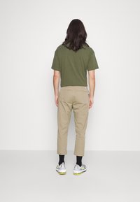 Olivově zelená košile s krátkým rukávem v kombinaci se světle béžovými zúženými kalhotami, které mají hladkou texturu a klasický střih, nošena s šedými teniskami.