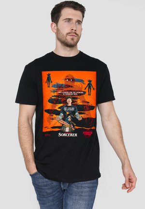 Mann trägt schwarzes T-Shirt mit buntem "Stranger Things" Kapitel Vier Zauberer Motiv, das Figuren und Text auf orangem Hintergrund zeigt.