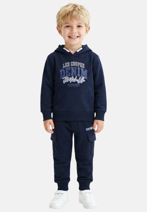 Garçon blond souriant, portant un sweat à capuche bleu marine "Lee Cooper Denim", un pantalon cargo assorti et des baskets blanches, debout devant un fond uni.