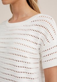 Vit stickad tröja med horisontella ränder och jämnt fördelade runda hål. Korta ärmar och en rund halsringning fulländar designen.