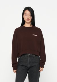 Felpa corta marrone in materiale morbido, con scollatura rotonda e maniche lunghe, decorata con un piccolo logo rosa "Tommy" sul petto. Abbinata a jeans scuri.