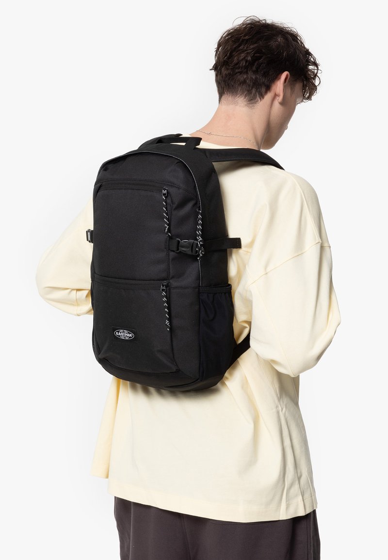 Mochila negra hecha de un tejido duradero, con bolsillos con cremallera, correas ajustables y un logo en la parte frontal. Diseño simple y funcional.