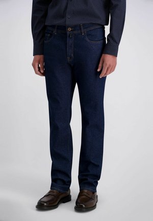 Ein Mann trägt dunkelblaue Jeans, braune Leder-Loafer und ein schwarzes Hemd mit kleinen weißen Punkten und steht vor einem einfarbigen Hintergrund.