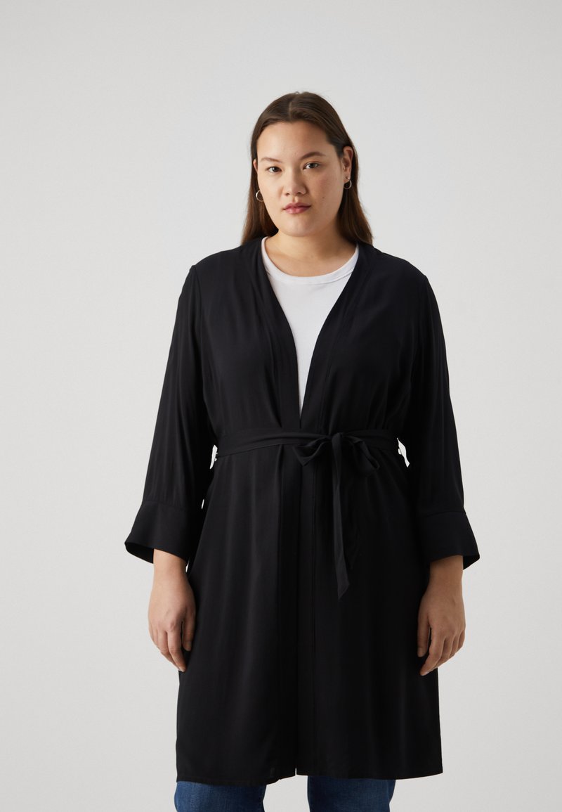 Robe noire à manches longues avec une taille à liens, en tissu lisse. Présente un col châle et des poignets larges, assortie à un haut blanc et un jean.
