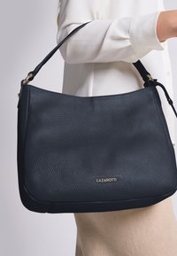 Schwarze Leder-Schultertasche mit strukturierter Oberfläche, ausgestattet mit einem goldenen Logo und einem einzelnen Schulterriemen. Oberteil und Hose in neutralen Farben sind teilweise sichtbar.