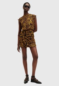 Robe imprimée léopard avec des épaules à volants, texture froncée et silhouette ajustée, associée à des chaussures noires à lanières et des lunettes de soleil.