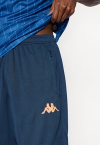 Tengerészkék sportnadrág texturált anyagból, oldalzsebbel és világos narancssárga logóval a combon, kék mintás toppal párosítva.