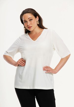 Frau trägt weißes V-Ausschnitt-Oberteil mit Ellbogenlangen Ärmeln und schwarze Hose, posiert mit Händen in der Hüfte vor einfachem Hintergrund.