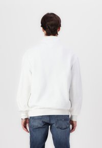 Felpa bianca dalla vestibilità casual, con polsini e orlo a costine, caratterizzata da un dettaglio a V sul retro, abbinata a jeans blu scuri.