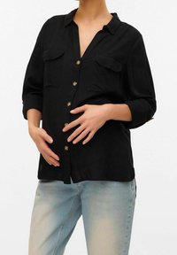 Camisa negra de botones con dos bolsillos en el pecho, hecha de tejido ligero. Presenta mangas enrolladas y botones en tono dorado. Combinada con jeans de color azul claro.
