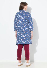 Blautöniger Tunika mit floralen Muster und asymmetrischem Saum, kombiniert mit burgundfarbenen Leggings mit Spitze an den Knöcheln und weißen Schuhen.