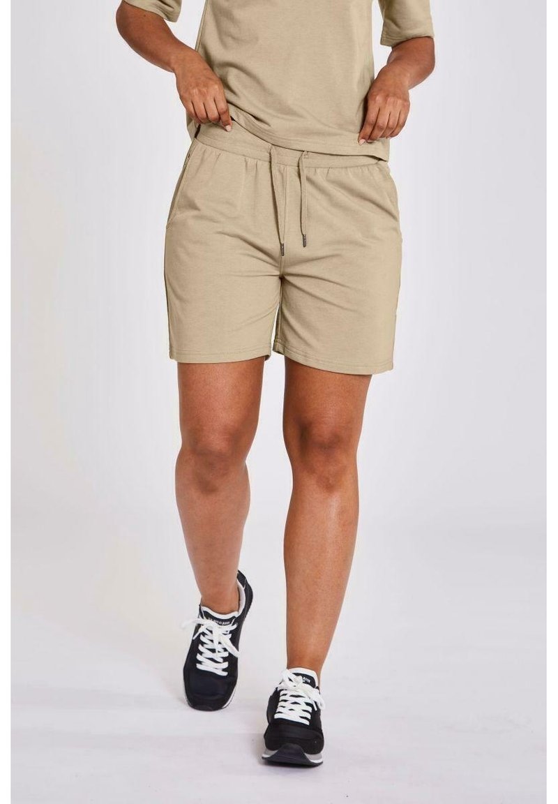 Person i beige shorts och matchande t-shirt, justerar midjebandet, svarta och vita sneakers, står mot en enfärgad bakgrund.