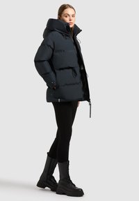 Puffer jacket in dark navy, featuring a high collar, drawstrings, and zip closure. Paired with black leggings and mid-calf lace-up boots.