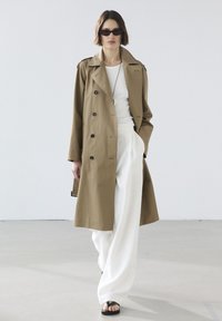 Khakifarbener Trenchcoat mit doppelter Knopfleiste, getragen über einem weißen Oberteil und weit geschnittenen weißen Hosen. Schwarze Sonnenbrillen runden das Outfit ab.