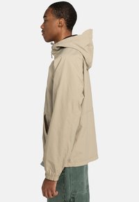 Beige hættejakke med let vævning, justerbar snørelukning i hætten og lynlåslommer foran. Enkel design og løs pasform.