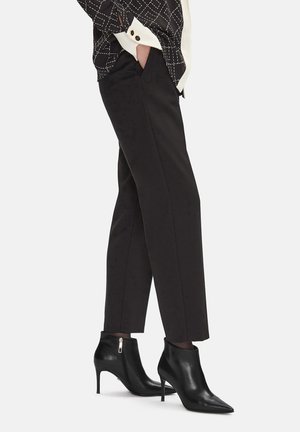 Kobieta ubrana w czarne, dopasowane spodnie i czarne, spiczaste botki na wysokim obcasie, z jedną ręką w kieszeni, stojąca na białym tle.