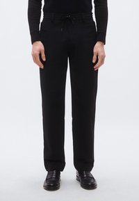 Čierne nohavice s voľným strihom, elastickým pásom, šnúrkou a hladkou textúrou látky. Štylizované s čiernymi topánkami a priliehavým čiernym vrškom.