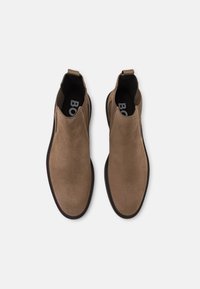 Pár hnědých semišových nazouvacích kotníkových bot s černými podrážkami a poutky, zobrazených z vrchu na bílém pozadí.