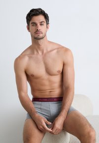 Fiatal férfi sötét hajjal, szürke boxer alsóneműt viselve, bordó derékszíjjal ül egy világos színű, texturált széken egy sima fal előtt.