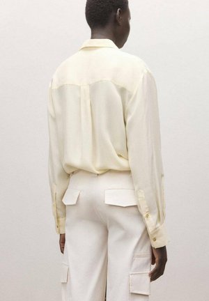 Persona que lleva una camisa de manga larga color crema claro metida dentro de pantalones cargo crema con bolsillos de solapa, de pie contra un fondo liso, de espaldas.