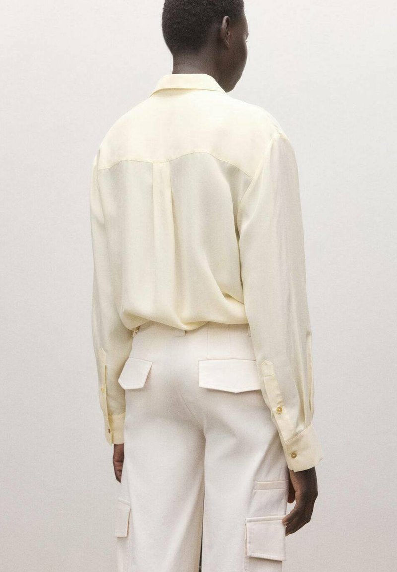 Personne portant une chemise à manches longues crème clair rentrée dans un pantalon cargo crème avec poches à rabat, debout devant un fond uni, de dos.