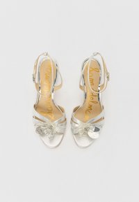 Silver high-heeled sandals with ankle straps, textured fabric, floral embellishment, and a cushioned insole with a gold logo.