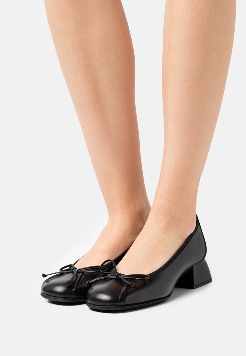 Femme portant des ballerines en cuir noir à petit talon avec un détail nœud, debout devant un fond clair uni.