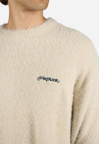 Beige kötött pulóver bolyhos textúrával, kerek nyakkivágással, és egy kis fekete hímzett logóval a bal mellen.