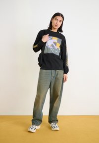 Schwarzes langärmeliges Shirt mit Grafiken und gelben sowie grauen Akzenten, kombiniert mit lockeren blauen Jeans und weißen Sneakers.