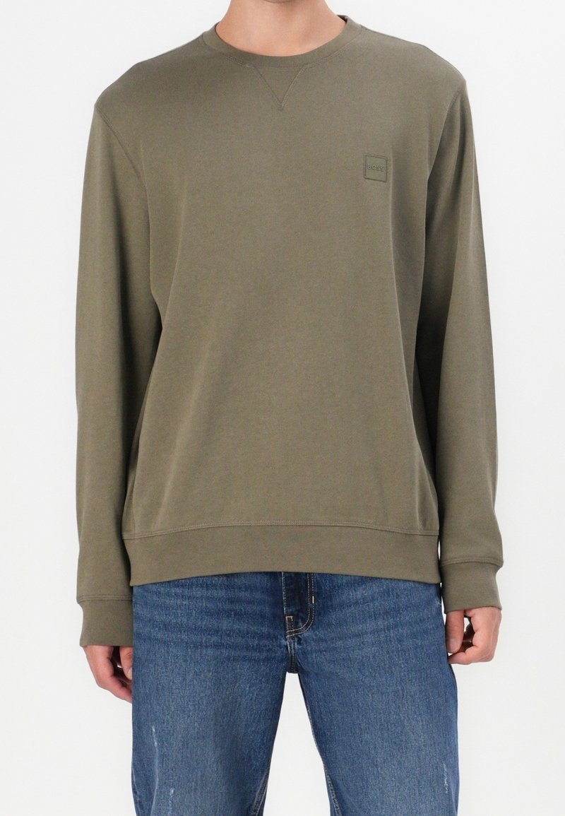 Homme portant un sweat-shirt à col rond et manches longues vert olive et un jean bleu, debout devant un fond blanc.