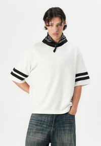 Hvid kortærmet skjorte med sorte striber på ærmerne, parret med et sort mønstret bandana. Båret med blå denimbukser.