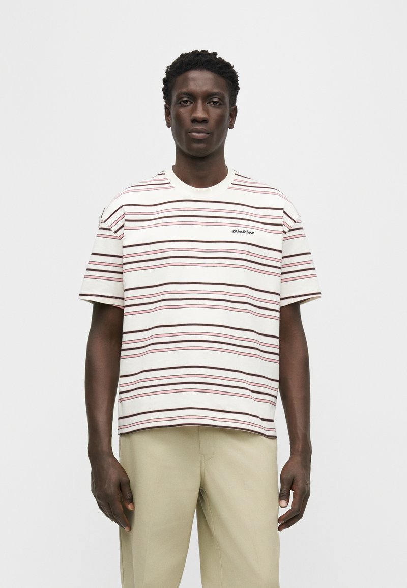 Hombre de pie que lleva una camiseta de manga corta color crema con rayas horizontales rojas y negras y pantalones beige sobre un fondo blanco.