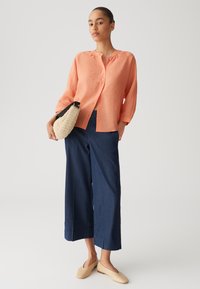 Camicia in lino corallo con maniche a tre quarti, abbinata a pantaloni in denim scuri a gamba larga e una borsa intrecciata con tracolla nera.