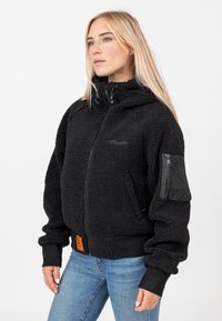 Veste en polaire noire à fermeture éclair, col montant et poignets côtelés. Présente une poche zippée sur la manche gauche et une étiquette orange en bas.