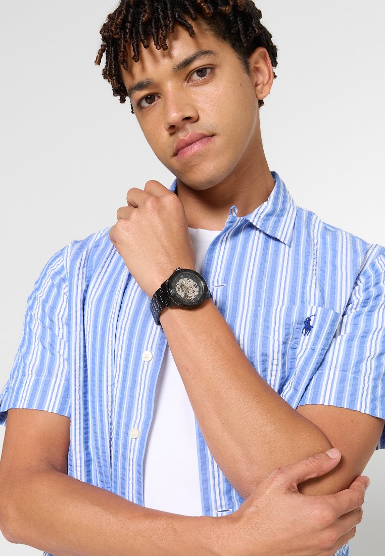 Orologio da polso nero con quadrante trasparente che mostra ingranaggi complessi, indossato su una mano. Modello che indossa una camicia a maniche corte a righe blu e bianche.